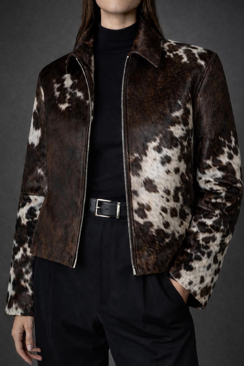 May include: A brown and white cowhide jacket with a zip-up front and a collar. The jacket is worn over a black turtleneck and paired with black pants and a black belt.