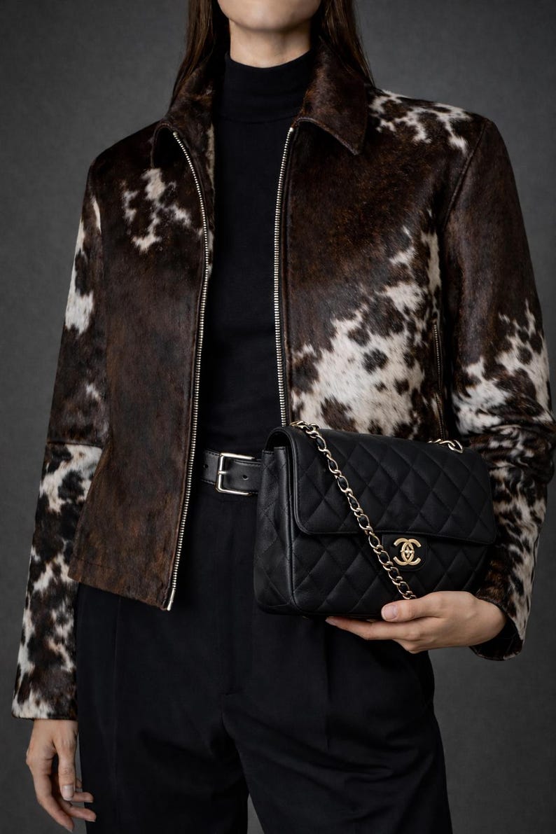 May include: A brown and white cowhide jacket with a zipper closure. The jacket is paired with a black turtleneck, black pants, and a black quilted handbag with a gold chain strap and logo.