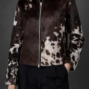 May include: A brown and white cowhide jacket with a zip-up front. The jacket has a collar and is paired with black pants. The cowhide pattern is a mix of dark brown and white spots. The jacket is a stylish outerwear option.