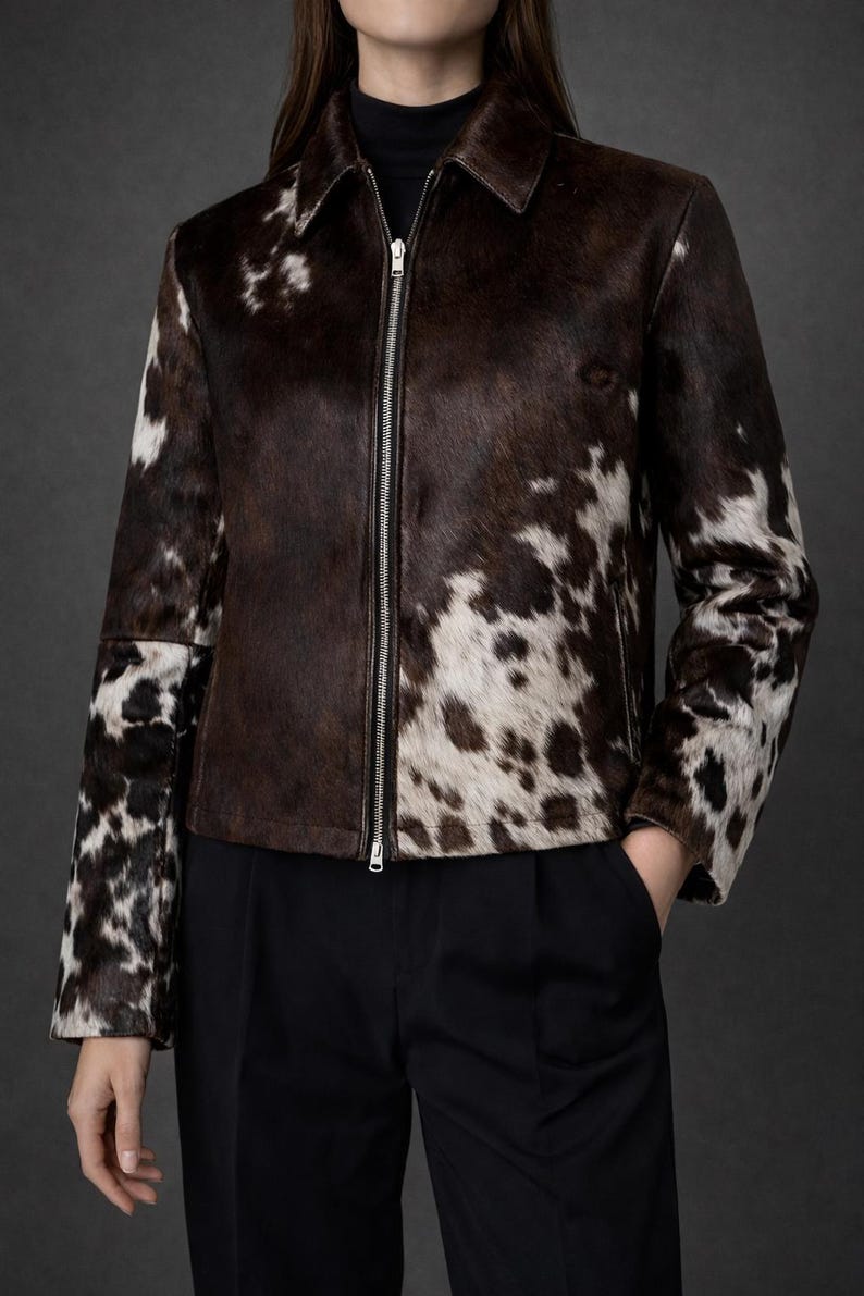 May include: A brown and white cowhide jacket with a zip-up front. The jacket has a collar and is paired with black pants. The cowhide pattern is a mix of dark brown and white spots. The jacket is a stylish outerwear option.