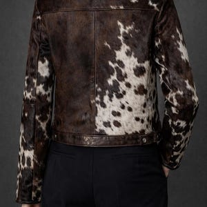 May include: A brown and white cowhide jacket with a classic collar and button closure. The jacket features a unique spotted pattern and is paired with black pants. The back view showcases the jacket's design.