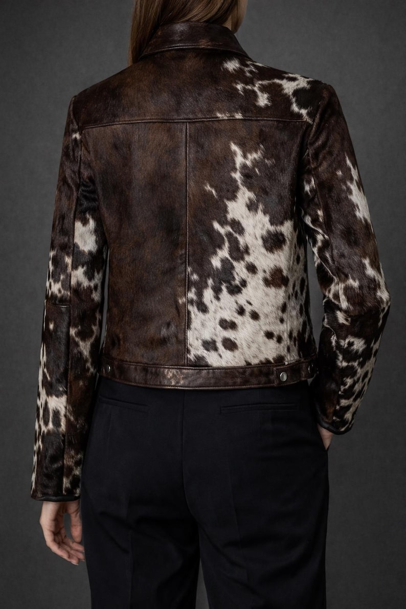 May include: A brown and white cowhide jacket with a classic collar and button closure. The jacket features a unique spotted pattern and is paired with black pants. The back view showcases the jacket's design.