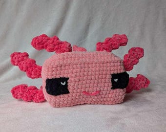 May include: A handmade pink crochet axolotl plushie with black eyes and pink gills. The plushie has a smiling mouth and is made of soft, textured yarn. The axolotl is a cute and cuddly toy.