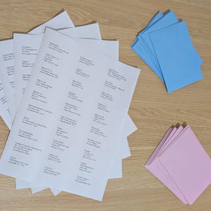 Puede incluir: Hojas blancas con etiquetas de direcciones impresas, junto con pilas de sobres azul claro y rosa, dispuestas sobre una superficie de madera. Las etiquetas están listas para enviar.