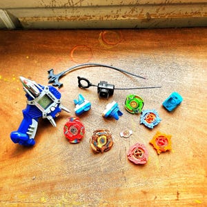 May include: A collection of colorful spinning top toys and a blue and white launcher with a digital display. The tops are various colors, including red, green, and orange. The toys are arranged on a wooden surface.