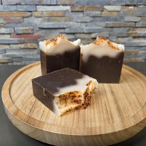 May include: Three handmade soaps with a layered design, presented on a wooden tray. The soaps have a dark brown base with a white top and a textured, light brown edge. The tray is round and made of light-coloured wood.
