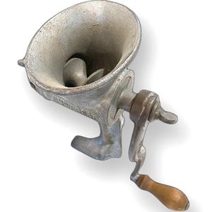 May include: A vintage, cast-iron meat grinder with a silver finish. The grinder features a large hopper, a hand crank with a wooden handle, and a base for stability. The design is classic and functional, showcasing the craftsmanship of a bygone era.