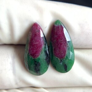 Peut inclure: Deux pierres précieuses en forme de goutte d'eau, avec un centre rouge profond et une couche extérieure verte. Les gemmes sont facettées, captant la lumière. Les pierres sont présentées sur un fond blanc, mettant en valeur leurs couleurs et textures naturelles. Ces gemmes sont probablement destinées à la joaillerie.