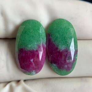 Könnte beinhalten: Zwei ovalförmige Edelsteine mit Facettenschliff. Jeder Stein ist in zwei Abschnitte unterteilt: eine leuchtend grüne obere Hälfte und eine tiefmagentafarbene untere Hälfte. Die Edelsteine werden vor einem dunklen Hintergrund präsentiert.