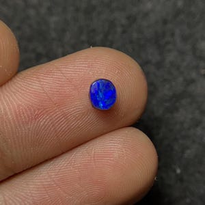 Op de afbeelding: Een kleine, ovaalvormige, levendige blauwe opaal edelsteen. De edelsteen vertoont een boeiend kleurenspel, met flitsen van heldere blauwe tinten. De opaal wordt tussen de vingers gehouden, wat de grootte en schittering laat zien.