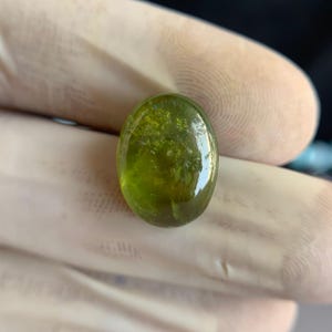 Op de afbeelding: Een ovale, doorschijnende edelsteen met een levendige groene tint. Het gepolijste oppervlak reflecteert licht en onthult interne insluitsels en een subtiele, natuurlijke textuur. De edelsteen wordt in een gehandschoende hand gehouden, wat de helderheid en kleur benadrukt.