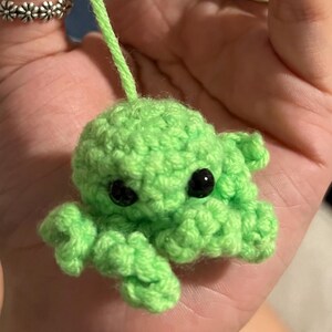 May include: A small, handmade crochet octopus in bright green. The octopus has black safety eyes and a green yarn loop for hanging. The tentacles are curled, and the body is round. The octopus is held in a hand.