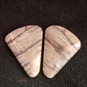 Puede incluir: Dos piedras pulidas de forma triangular con una superficie lisa de color base beige claro y patrones lineales de color marrón oscuro, similares a la veta de la madera, que corren diagonalmente a través de cada pieza. Son simétricas en tamaño y forma.