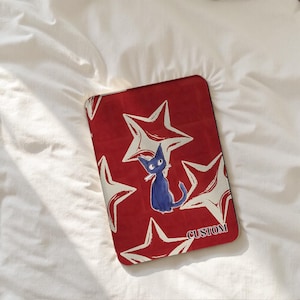 Może przedstawiać: Czerwone etui na tablet z wzorem białych gwiazd i ilustracją niebieskiego kota. Słowo "CUSTOM" jest wydrukowane na dole po prawej stronie. Etui jest prostokątne i wydaje się być wykonane z twardego materiału.