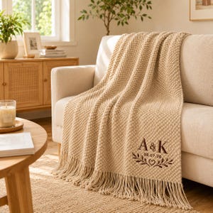 May include: A beige textured knit throw blanket featuring a fringe edge and dark brown embroidery. The embroidery displays the letters A & K, the date 08.07.26, and a decorative branch motif with a small heart centred at the bottom.