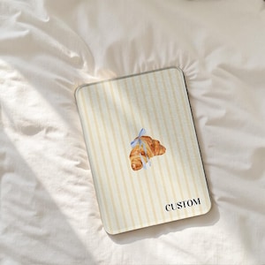 May include: Rectangular case featuring a cream and yellow striped design. A watercolour illustration of a croissant tied with a blue ribbon is on the front. The word "CUSTOM" is printed in black on the bottom right corner.