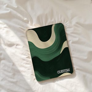 Puede incluir: Una funda de teléfono rectangular y personalizada con un patrón ondulado en tonos verdes y crema. La palabra "CUSTOM" está impresa en blanco en la esquina inferior derecha. La funda está sobre una sábana blanca.