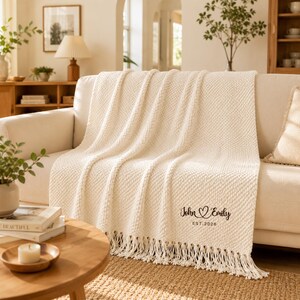 May include: A cream-coloured knit throw blanket featuring a textured pattern and fringe trim along the bottom edge. The blanket is personalised with the black text "John ♡ Emily" and "EST. 2026" embroidered in a script font on the lower right corner.