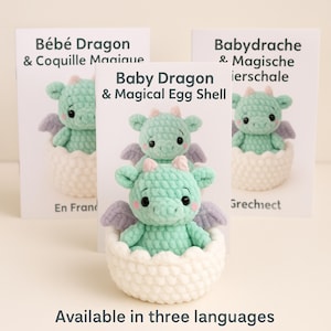 Op de afbeelding: Een gehaakte babydraak in een witte eierschaal. De draak is lichtgroen met paarse vleugels en kleine hoorns. De afbeelding bevat drie kaarten met tekst in verschillende talen.