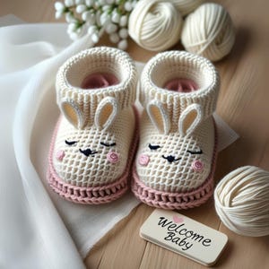 Può includere: Un paio di scarpine da neonato all'uncinetto fatte a mano, color crema, con facce di coniglietto e dettagli rosa. Le scarpine hanno un polsino a maglia e una base rosa. Un piccolo cartello dice "Welcome Baby".