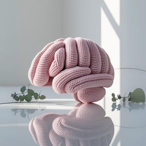 May include: A pink, crocheted brain model sits on a reflective surface. The brain is detailed with visible lobes and textures, and is the focal point of the image. Green sprigs of foliage are placed near the brain.