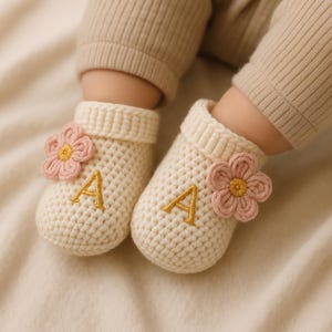 以下が含まれることがあります： クリーム色のベビー用かぎ針編みブーツ。ピンクの花と金色の刺繍文字「A」が特徴です。ベージュのニットパンツを履いた赤ちゃんの足元にブーツが写っています。