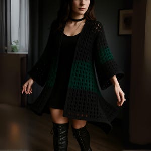 Peut inclure: Un cardigan en crochet noir et vert à devant ouvert avec des manches cloche. Le cardigan présente un motif géométrique et un dégradé vert sur la moitié inférieure. La modèle porte une robe noire et des bottes noires.