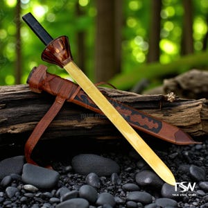 May include: A decorative sword with a light gold blade, a brown and black handle, and a brown leather sheath. The sword is resting on a piece of wood with a background of green foliage and black stones. The text "TRUE STEEL WORK" is visible.