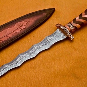 May include: A decorative dagger with a wavy, patterned blade and a detailed handle. The handle features a brown and black chevron design. The dagger is accompanied by a brown leather sheath with a leaf design. The background is a solid orange color.