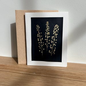 May include: A greeting card featuring a black background with three sprigs of dried flowers. The card is framed in white and placed in front of a brown envelope. The card and envelope are on a wooden surface.