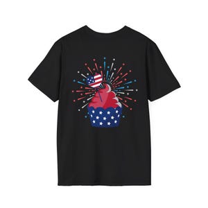 May include: Black t-shirt featuring a patriotic cupcake design. The cupcake has a red frosting top and a blue base decorated with white stars. A small American flag is placed on top, with a burst of red, white, and blue fireworks.