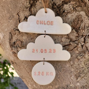 Könnte beinhalten: Ein Set aus drei Keramikornamenten: zwei Wolken und ein Kreis. Die obere Wolke trägt den Namen "CHLOE" in Rosa. Die mittlere Wolke zeigt das Datum "21.03.23". Der untere Kreis zeigt "3.20 KG". Aufgehängt an einer braunen Schnur.