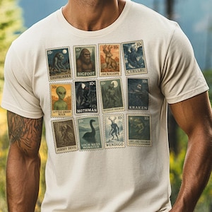Pode incluir: Camiseta bege com uma coleção de selos postais em estilo vintage com ilustrações de criptídeos. Os selos retratam criaturas como Bigfoot, Chupacabra e o Monstro do Lago Ness. Cada selo tem um valor de "10c". A camiseta tem gola redonda.