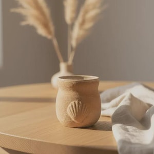 Peut inclure: Une petite tasse en céramique beige, ronde, avec une surface texturée et un motif en relief représentant un coquillage sur le devant. La tasse présente une large ouverture et une finition mate, affichant une teinte naturelle et terreuse.
