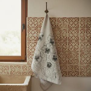 Puede incluir: Un paño de cocina beige colgado de un gancho de pared, con una ilustración de líneas negras de flores de pensamiento. La tela tiene una textura tejida natural y el diseño floral cubre el centro con pétalos y hojas detallados.