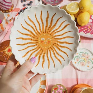 Puede incluir: Plato de cerámica blanco con borde festoneado, con un diseño de sol en naranja y amarillo. El sol tiene una cara sonriente con corazones por ojos. Otros platos, tazas y limones en el fondo.