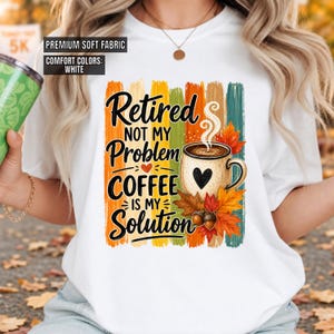 May include: A white short-sleeve t-shirt featuring a graphic print with vertical brush strokes in orange, yellow, green, and teal. The text reads "Retired Not My Problem Coffee Is My Solution" alongside a coffee mug, autumn leaves, and acorns.
