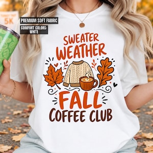 May include: A white t-shirt featuring a graphic with the text "SWEATER WEATHER FALL COFFEE CLUB" in orange and brown. The design includes a beige cable-knit sweater, a brown mug of coffee, and orange autumn leaves. Text boxes read "PREMIUM SOFT FABRIC" and "COMFORT COLORS: WHITE".
