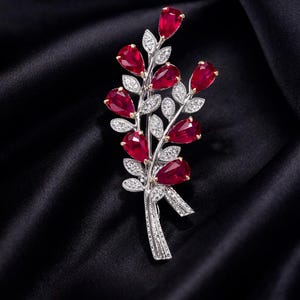 Könnte beinhalten: Eine silberfarbene Brosche in floralem Design mit tropfenförmigen roten Edelsteinen und kleinen weißen Edelsteinen. Die Brosche ist vor einem schwarzen Satinhintergrund platziert, der die Details hervorhebt.