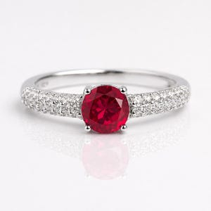 Peut inclure: Une bague en argent avec une grosse pierre rouge foncé et ronde au centre. La bande est incrustée de petits diamants transparents. La bague est mise en valeur sur un fond blanc, soulignant son éclat et son design.