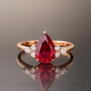 May include: A rose gold ring featuring a central pear-shaped deep red gemstone held by prongs. The band is accented on each side by a cluster of three small, clear, round-cut stones. The ring is displayed against a reflective surface.