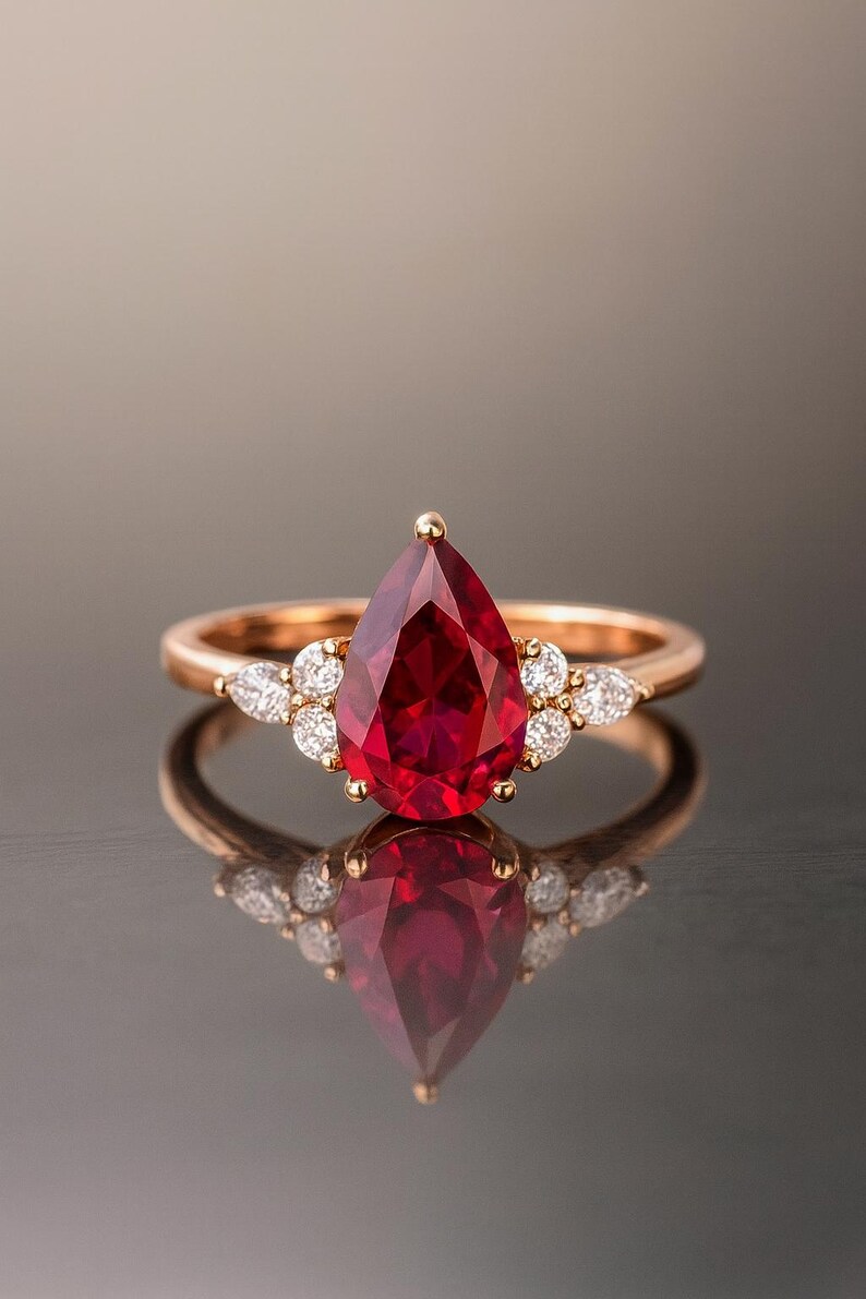 May include: A rose gold ring featuring a central pear-shaped deep red gemstone held by prongs. The band is accented on each side by a cluster of three small, clear, round-cut stones. The ring is displayed against a reflective surface.