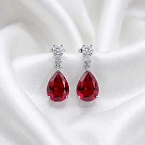 Puede incluir: Un par de pendientes colgantes de tono plateado con una gran piedra roja facetada en forma de pera suspendida de un engaste vertical. Cada pendiente incluye dos pequeñas piedras redondas, claras y de talla brillante sobre la gema principal.