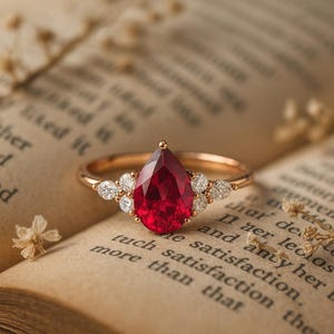 May include: A gold ring featuring a central pear-shaped red gemstone flanked by three small round white stones on each side. The ring is positioned on the open pages of a vintage book with visible printed text and small dried floral sprigs.