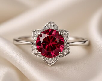 May include: A silver ring with a flower-shaped setting, showcasing a large, round, deep red gemstone. The petals are adorned with smaller, sparkling white stones. The ring is set against a soft, white background.