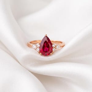 May include: A rose gold ring featuring a central pear-shaped red gemstone. The setting includes three small round clear stones on each side of the main gem, arranged in a cluster along the thin, polished rose gold band.