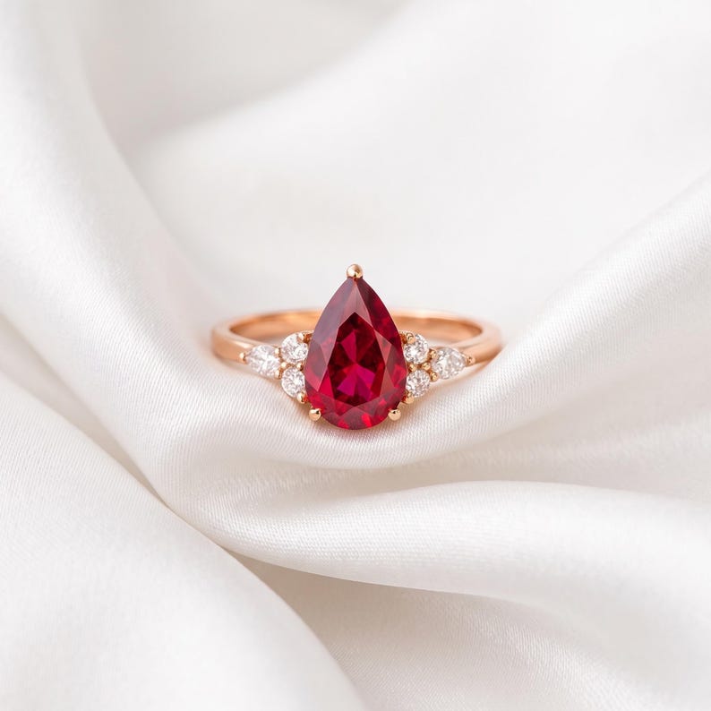 May include: A rose gold ring featuring a central pear-shaped red gemstone. The setting includes three small round clear stones on each side of the main gem, arranged in a cluster along the thin, polished rose gold band.