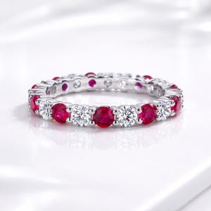 Puede incluir: Una alianza de eternidad en plata con un patrón alterno de rubíes rojos vibrantes y diamantes blancos transparentes de talla redonda, montados en engaste de garras compartidas. El anillo muestra un diseño circular continuo con garras de metal pulido.