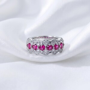 May include: A silver-toned ring featuring a wavy band design set with a row of six round, vibrant pinkish-red gemstones. The band is further embellished with numerous small, clear, sparkling stones arranged in a pavé setting.