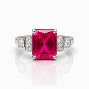 May include: A silver ring featuring a large, rectangular, vibrant pink gemstone as the centerpiece. The ring is flanked by smaller, clear gemstones on each side, set against a white background. The ring's design is elegant and eye-catching.